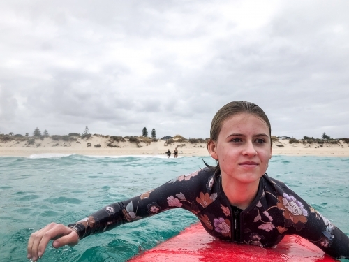 Close up of Teenage girl paddling red surfboard on ocean with beach in background - Australian Stock Image