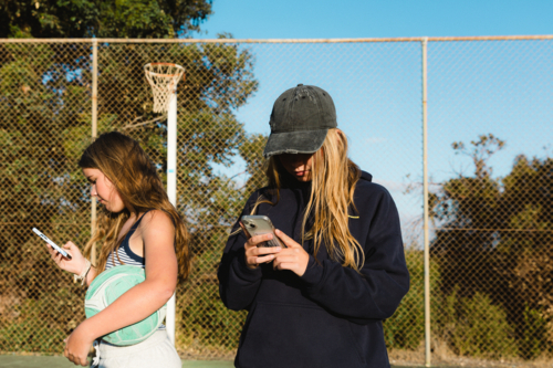 Close up of two teenage girls on smart phones holding ball at outdoor court - Australian Stock Image