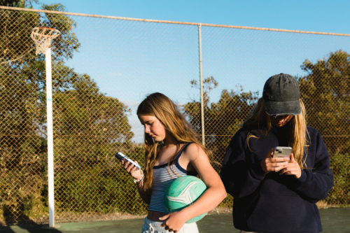 Close up of two teenage girls on smart phones whilst holding netball at outdoor court - Australian Stock Image