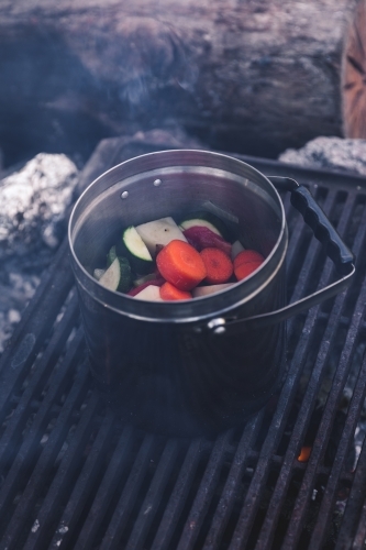 Close up of vegetables cooking in steel pot on the grill on a smoking campfire - Australian Stock Image