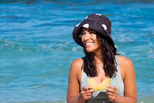 Close up of Young woman wearing full brim hat and eating rockmelon on the beach on clear sunny day - Australian Stock Image