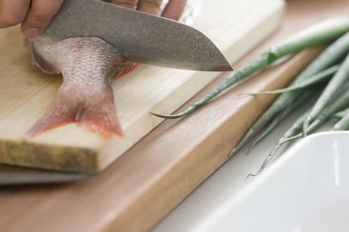 Close up photo of a red fish being sliced by a grey knife on wooden chop board