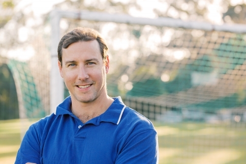 Close up photo of a smiling man wearing blue shirt on a sunny day with goal in the background - Australian Stock Image