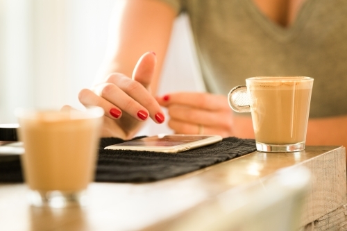 Close up photo of a woman's hand with red nails using her cellphone with two cups of coffee - Australian Stock Image