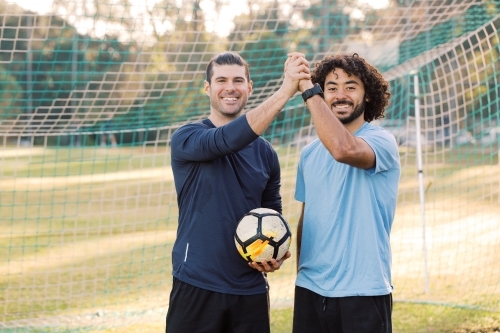 Close up photo of two male friends standing on the field holding and raising their hands with a ball - Australian Stock Image