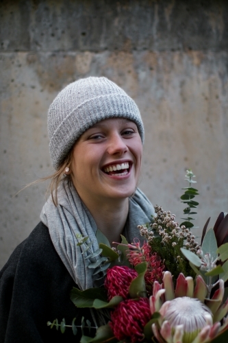 Close up portrait of young girl wearing a beanie and scarf smiling whilst holding a floral bouquet - Australian Stock Image