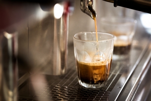 Close up shot of a coffee pouring from a machine into a glass cup on a metal counter - Australian Stock Image