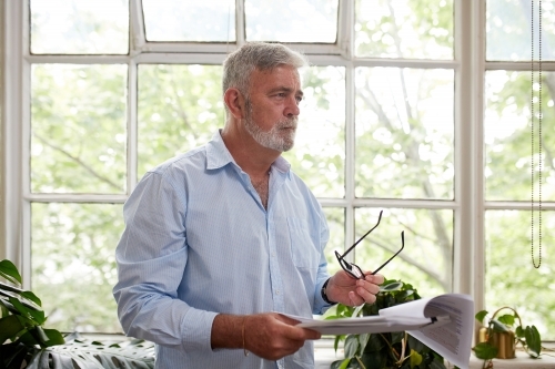Close up shot of a grey-haired adult standing behind a window holding his eye glasses and files - Australian Stock Image