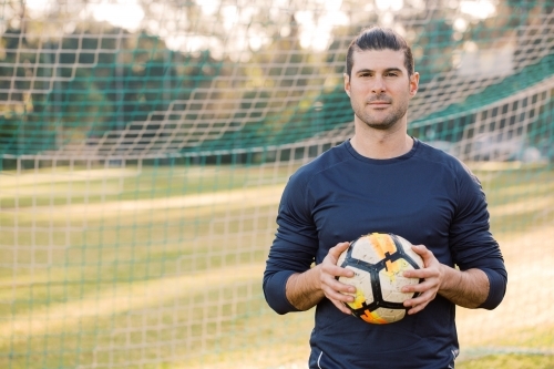 Close up shot of a man standing on the soccer filed, holding a soccer ball with a net behind - Australian Stock Image