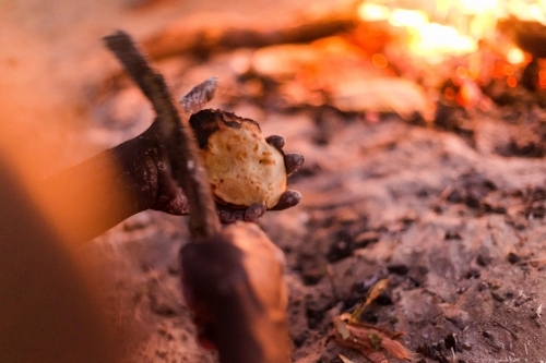 Close up shot of a person with dirty hands holding a jonny cake and a branch  with blurry flames - Australian Stock Image
