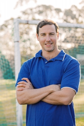 close up shot of a smiling man wearing blue shirt with arms crossed with goal in the background - Australian Stock Image