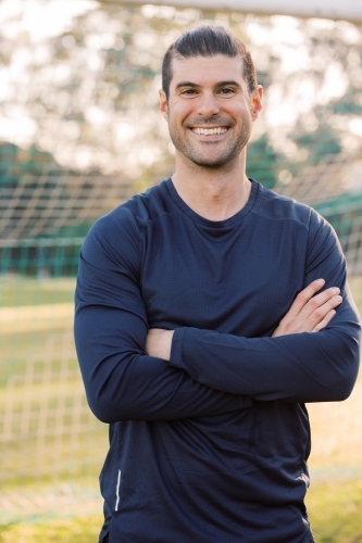Close up shot of a smiling young man, standing on the  field with crossed hands, wearing blue shirt - Australian Stock Image