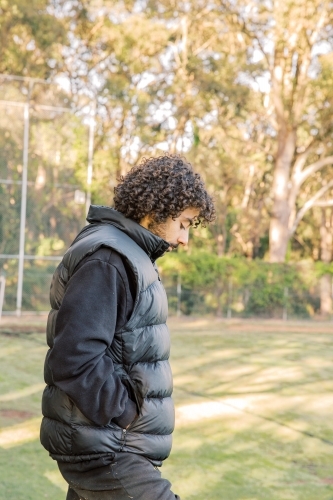 Side view shot of a young men standing on the field with a curly hair looking down with a black vest - Australian Stock Image