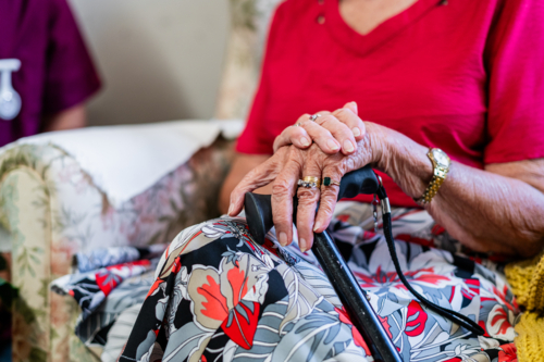Closeup of an elderly womans hand clasps together holding a cane while sitting on patterned armchair - Australian Stock Image