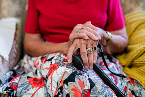 Closeup of an elderly womans hand clasps together holding a cane while sitting on patterned armchair - Australian Stock Image