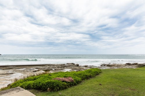 Cloudy skies over beach with ocean waves behind grass - Australian Stock Image