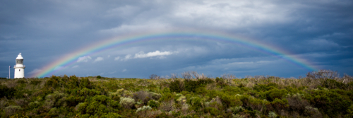 Coastal lighthouse beneath a rainbow with storm clouds in the background. - Australian Stock Image