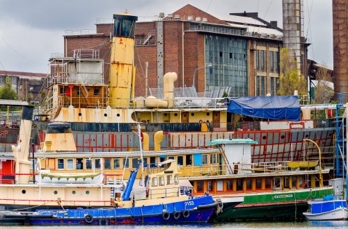 Colourful derelict boats and run down industrial building in background with smashed windows - Australian Stock Image