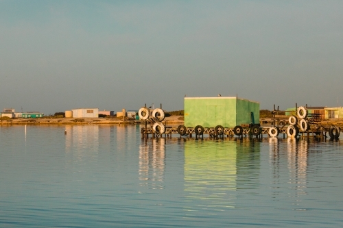 Colourful fishing shacks and shed with wooden jetty and old tyres, reflected in calm water