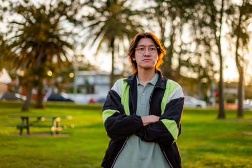 Confident young man poses with crossed arms in a green park at sunset, surrounded by palm trees - Australian Stock Image