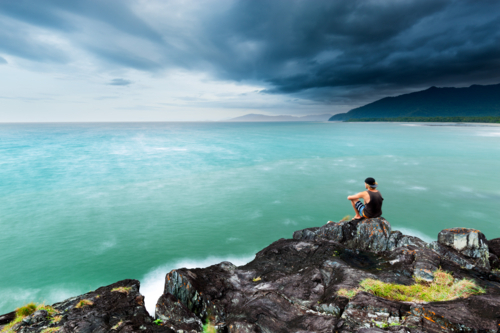 Contemplative young man sitting on a rock, looking at stormy ocean seascape and tropical coastline - Australian Stock Image