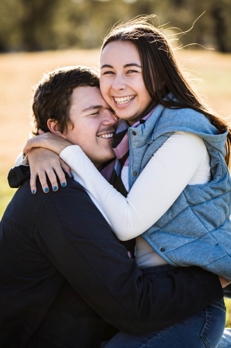 Couple cuddling with girlfriend sitting on boyfriend's lap with arms around neck laughing - Australian Stock Image
