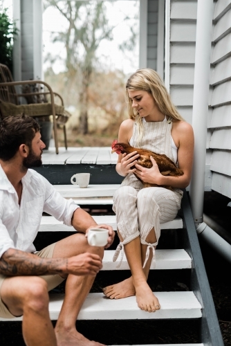 Couple having a cup of coffee on steps of verandah, with woman holding a chicken. - Australian Stock Image