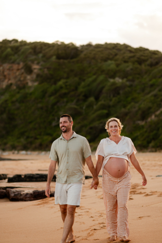 Couple holding hands walking on beach at sunset, pregnant woman in relaxed clothing - Australian Stock Image