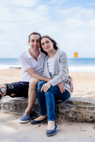 Couple relaxes on a rocky shoreline, smiling warmly against a scenic beach backdrop - Australian Stock Image