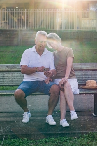 couple sitting on park bench, using phone - Australian Stock Image