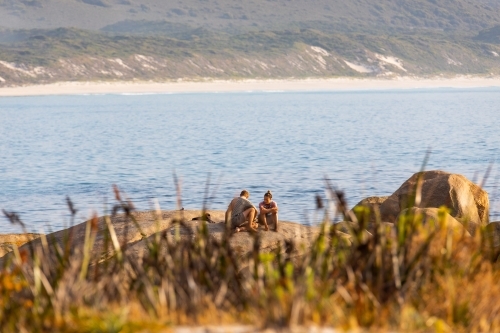 couple sitting on rock by the sea seen through blurred grasses - Australian Stock Image