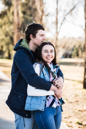 Couple standing with man's arms wrapped around woman holding hands looking off into distance - Australian Stock Image