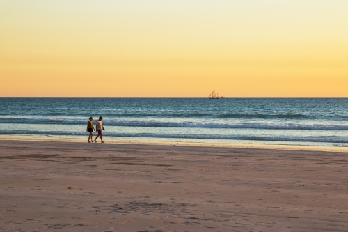 Couple walking along beach at sunset with boat sailing by - Australian Stock Image