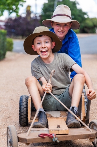 Cousins racing billy cart down driveway together - Australian Stock Image