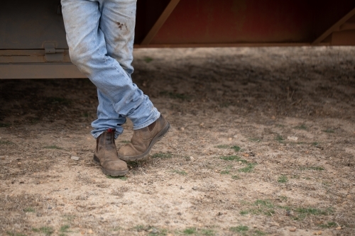 crossed legs of man standing wearing dirty jeans and work boots - Australian Stock Image