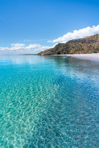 Crystal clear turquoise sea water along pebble beach with coastal cliffs under blue sky - Australian Stock Image