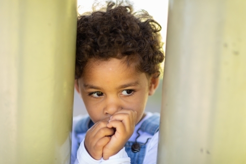curly-headed child sucking thumb and hiding - Australian Stock Image