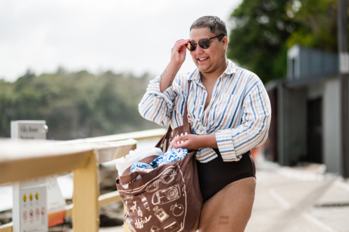 Curvy woman standing on coastal path wearing sunglasses carrying a brown beach bag on sunny day - Australian Stock Image