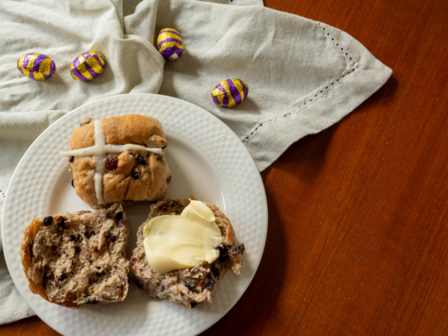Cut hot cross buns with butter on a white plate with linen napkins, foil easter eggs on wooden table - Australian Stock Image
