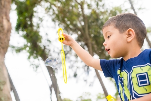 Cute mixed race 4 year old boy blows bubbles in a park by a lake - Australian Stock Image