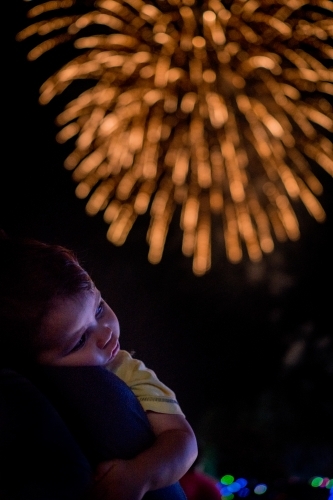 Cute mixed race baby boy cuddles his parent while new year's fireworks explode in background - Australian Stock Image