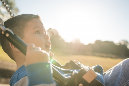 Cute mixed race three year old boy playing on a swing in a suburban playground - Australian Stock Image