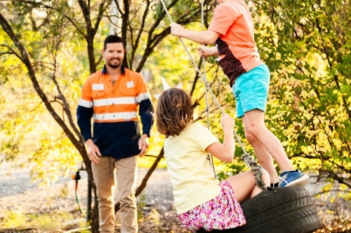 Dad playing with children in backyard on tyre swing in the afternoon - Australian Stock Image