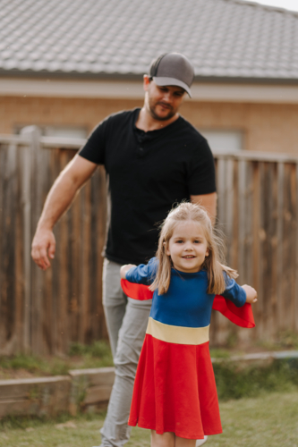 Dad playing with daughter in superhero costume in their yard. - Australian Stock Image