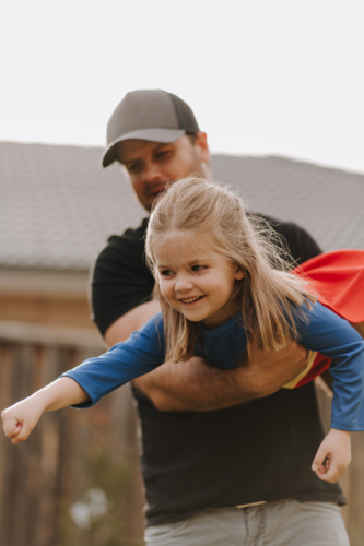 Dad simulating a flight for daughter in superhero costume. - Australian Stock Image