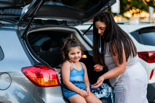 Daughter sits on the back of a car while mother talks to her in a parking area at dusk. - Australian Stock Image