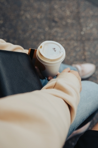 Detail of a young woman holding a coffee and phone waiting at a bus stop - Australian Stock Image