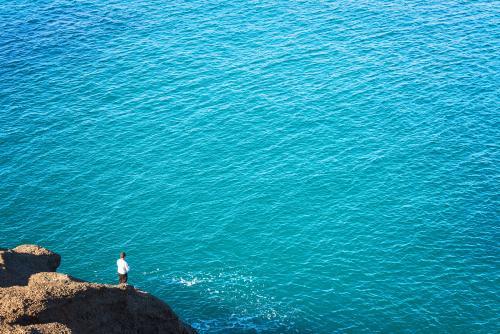 Distant person fishing off a rock beside clear blue sea waters