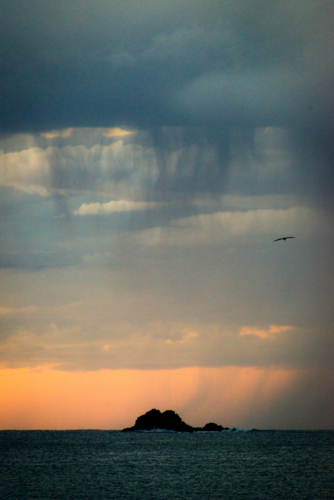 Distant rain falling over ocean at dusk with silhouette of rocky islet - Australian Stock Image
