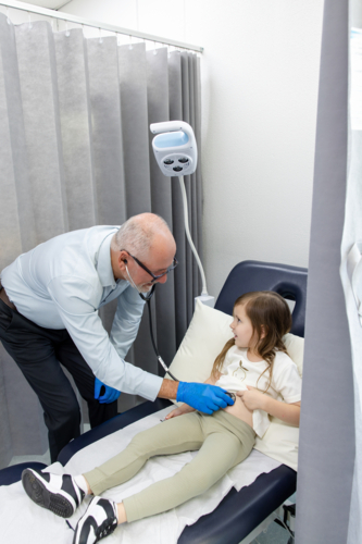 Doctor checking young girls tummy with mum standing carrying toddler son - Australian Stock Image
